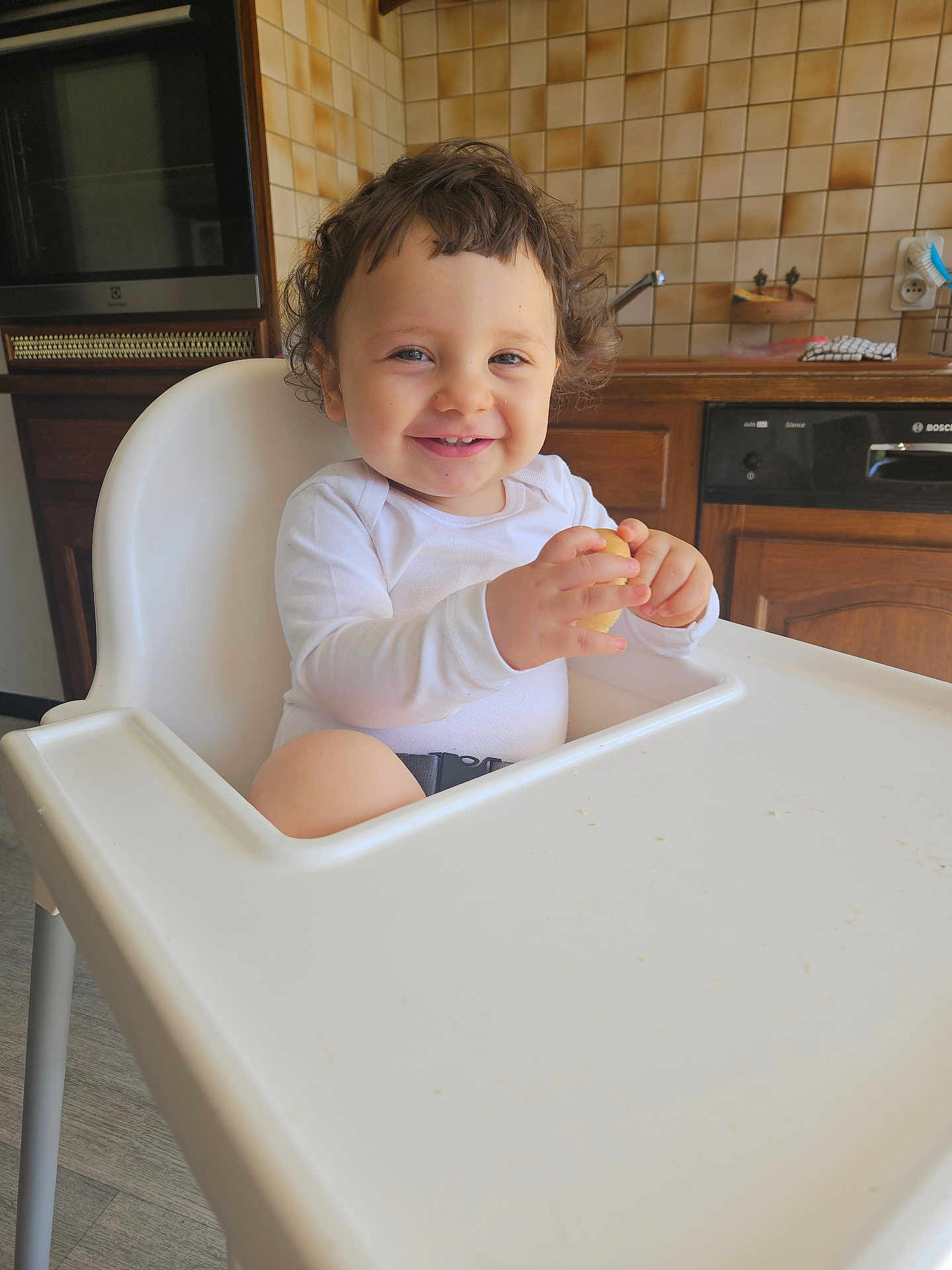Rael a rejoint le concours — aidez-le/la à gagner de superbes lots ! toddler, child, high_chair, smiling, curly_hair, white_clothing, kitchen, wooden_cabinets, tile_wall, snack, indoor, happy, baby, person, face, table, furniture, home, seat, food