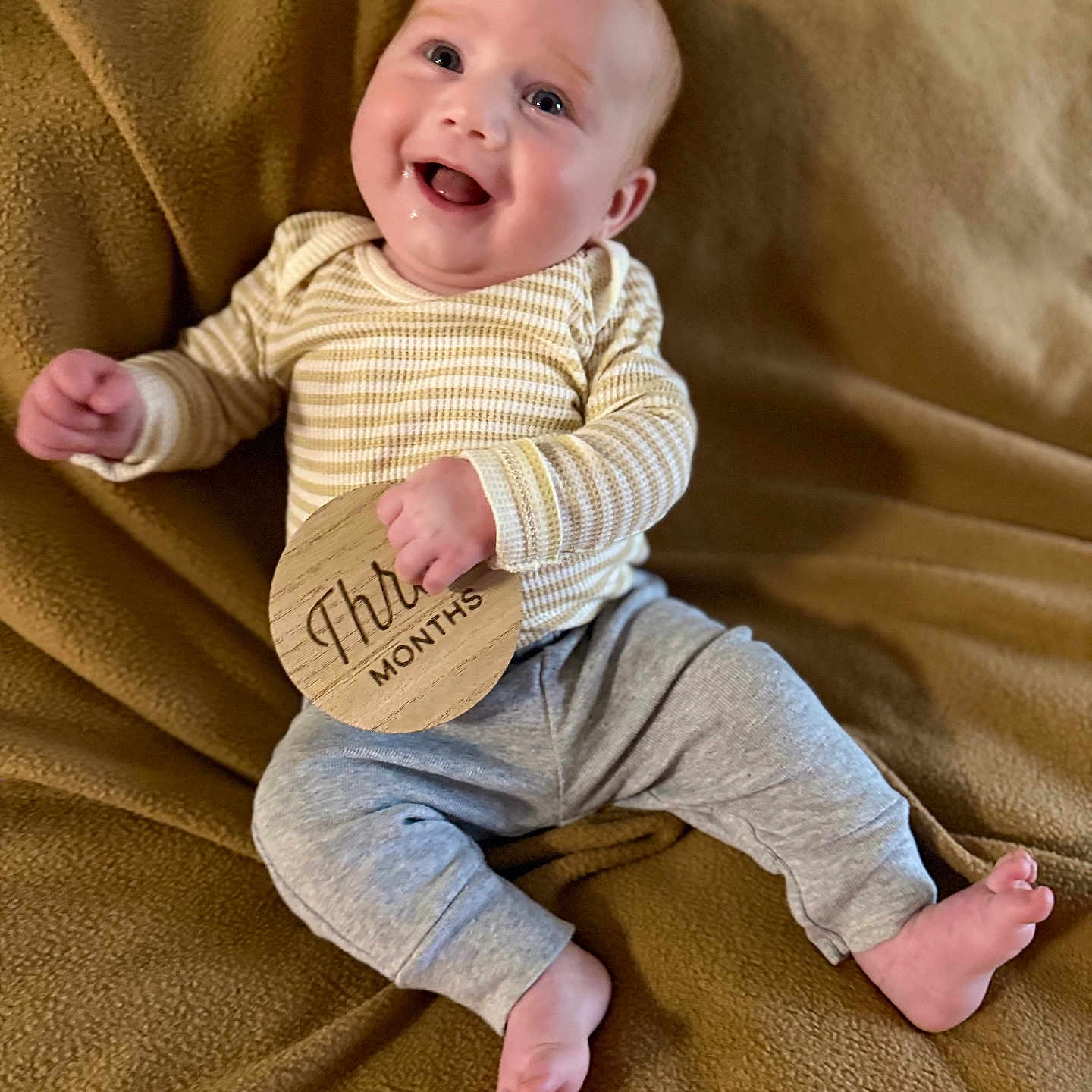 Ollie joined the competition — help win amazing prizes! baby, blanket, child, cozy, cute, face, gray_pants, happy, human, indoors, infant, lying_down, milestone, milestone_marker, portrait, small_feet, small_hands, smiling, striped_clothing, wooden_sign