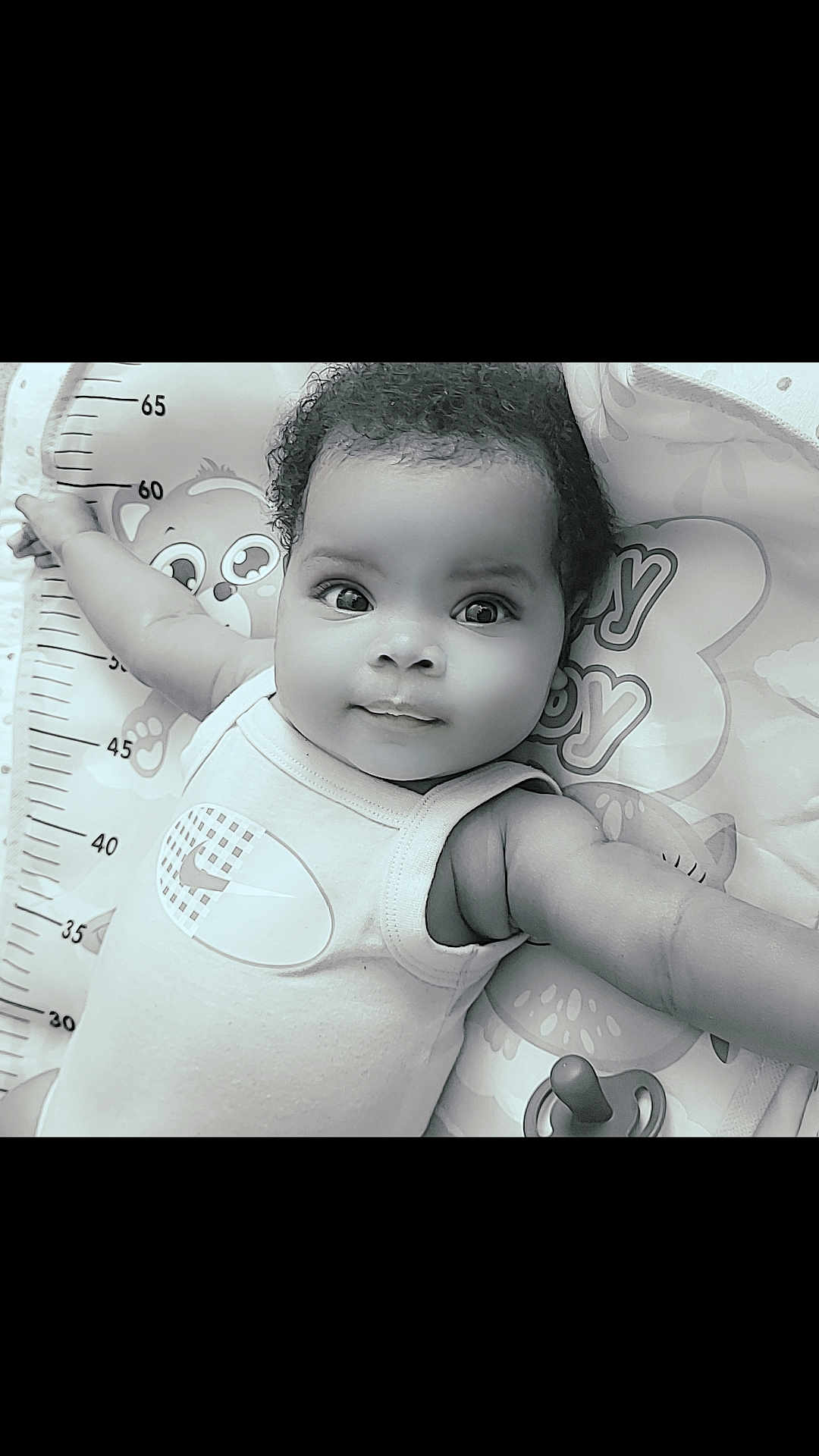 Kea'Ari is registered to the contest to win money with this photo: baby, infant, child, face, eyes, arms, curly_hair, smile, onesie, baby_mat, cartoon, measurement_scale, pacifier, cute, portrait, laying_down, indoors, soft_lighting, black_and_white, close_up