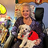 chair, cozy, decoration, dog, furniture, gray_hair, happy, home, indoor, jewelry, lamp, long_sleeves, pet, portrait, quilt, red_dress, seated, small_dog, smiling, woman