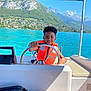 Ethan participe au concours pour gagner de l'argent avec cette photo : child, life_jacket, boat, steering_wheel, water, lake, mountains, outdoor, sunny, smiling, summer, vacation, nature, travel, recreation, blue_sky, greenery, adventure, person, leisure