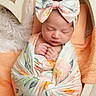 Jennie is registered to the contest to win money with this photo: newborn, baby, sleeping, swaddled, headband, bow, floral_pattern, blanket, cradle, cozy, infant, peaceful, wrapped, soft_texture, orange_towel, cute, portrait, indoors, child, resting