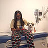 casual_clothing, clinic, companion, cute_dog, dog, focused, glasses, indoor, leisure, long_hair, medical_equipment, medical_exam_table, pajamas, pet, phone, red_vest, sitting, technology, white_wall, woman