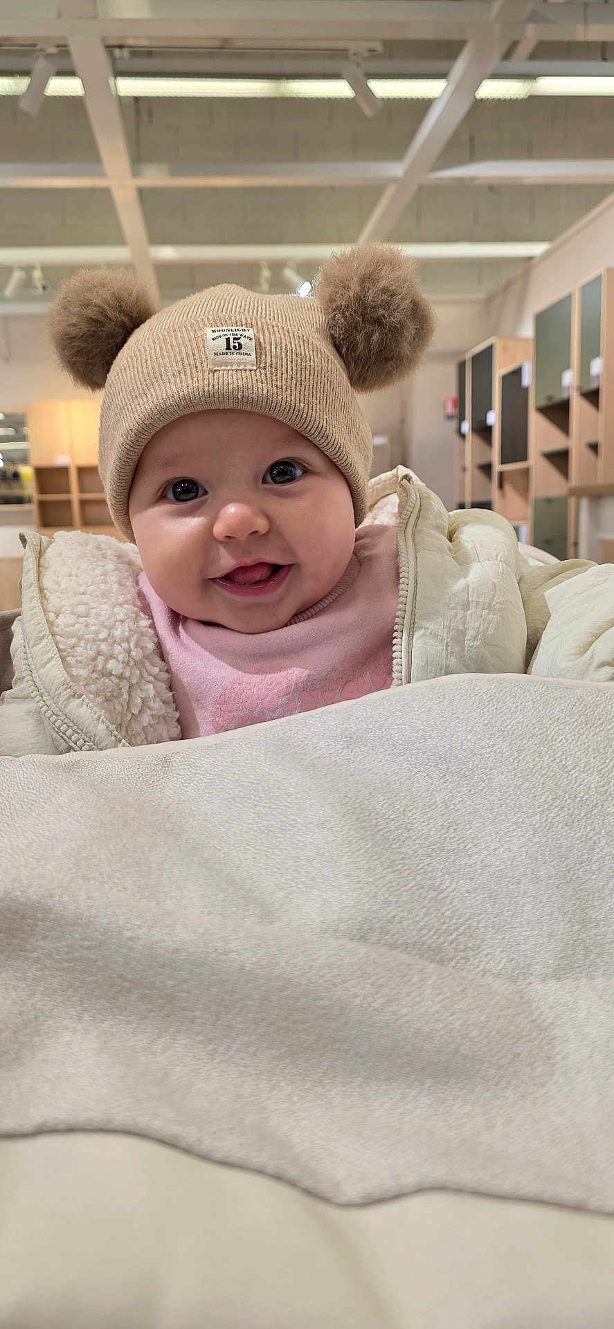 Anaya a rejoint le concours — aidez-le/la à gagner de superbes lots ! baby, child, hat, pom_poms, jacket, smile, tongue_out, indoor, cozy, cute, face, infant, warm_clothing, headwear, person, portrait, furniture, shelves, lighting, happy