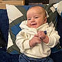 baby, child, smiling, white_hoodie, blue_jeans, pillow, blanket, indoor, couch, clapping, happy, cute, face, infant, person, seated, casual_clothing, home, furniture, relaxed