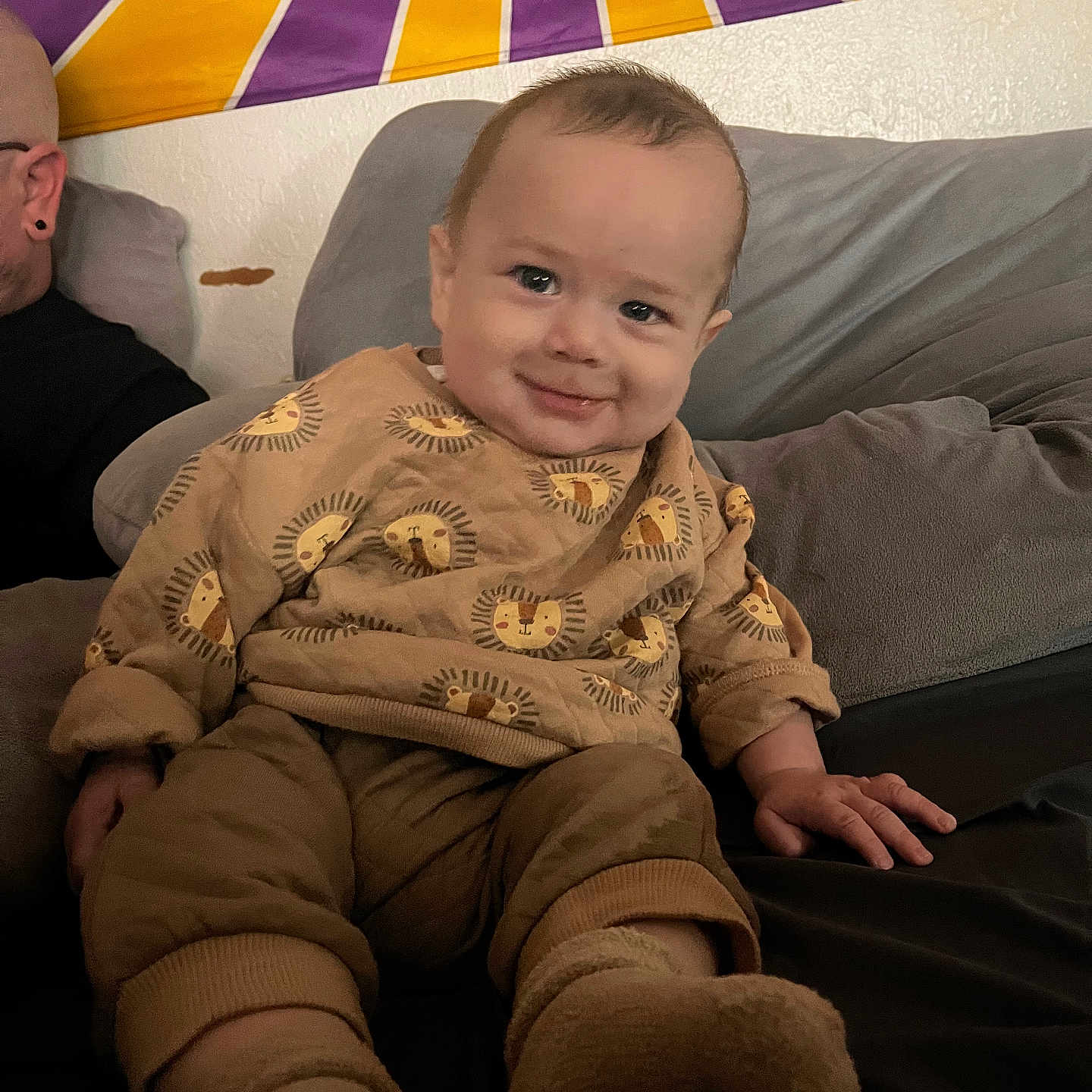 Angel L. B. joined the competition — help win amazing prizes! adult, baby, child, colorful_background, couch, cozy, earring, glasses, hand, indoor, lion_pattern, pajamas, person, pillow, relaxed, seated, smile, socks, tapestry, wall