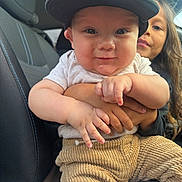 Gunner is registered to the contest to win money with this photo: baby, child, car_seat, clothing, cap, corduroy_pants, cuddling, cute, face, family, holding, infant, inside_car, person, portrait, smiling, sibling, toddler, young_child, window