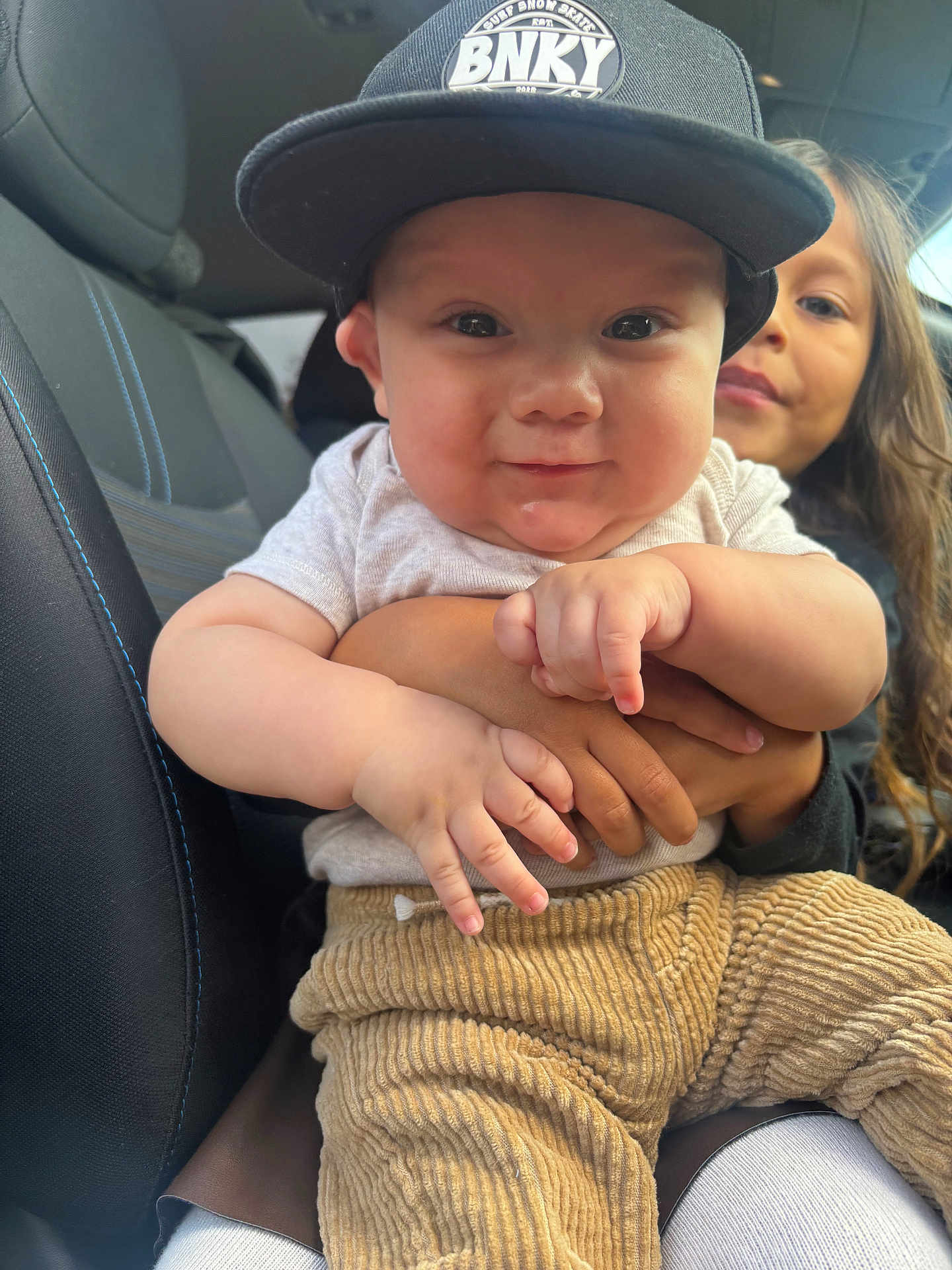 Gunner is registered to the contest to win money with this photo: baby, child, car_seat, clothing, cap, corduroy_pants, cuddling, cute, face, family, holding, infant, inside_car, person, portrait, smiling, sibling, toddler, young_child, window