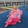 blue_surface, building, casual_clothing, child, curly_hair, daytime, fun, grass, happy, outdoor, person, pink_frog, play, playground, recreation, sandals, sculpture, sidewalk, smiling, young