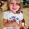 blonde_hair, casual_clothing, chair, child, classroom, daylight, desk, education, flower_graphic, happy, indoor, kid, missing_tooth, person, playful, school_supplies, smiling, table, tshirt, young_child