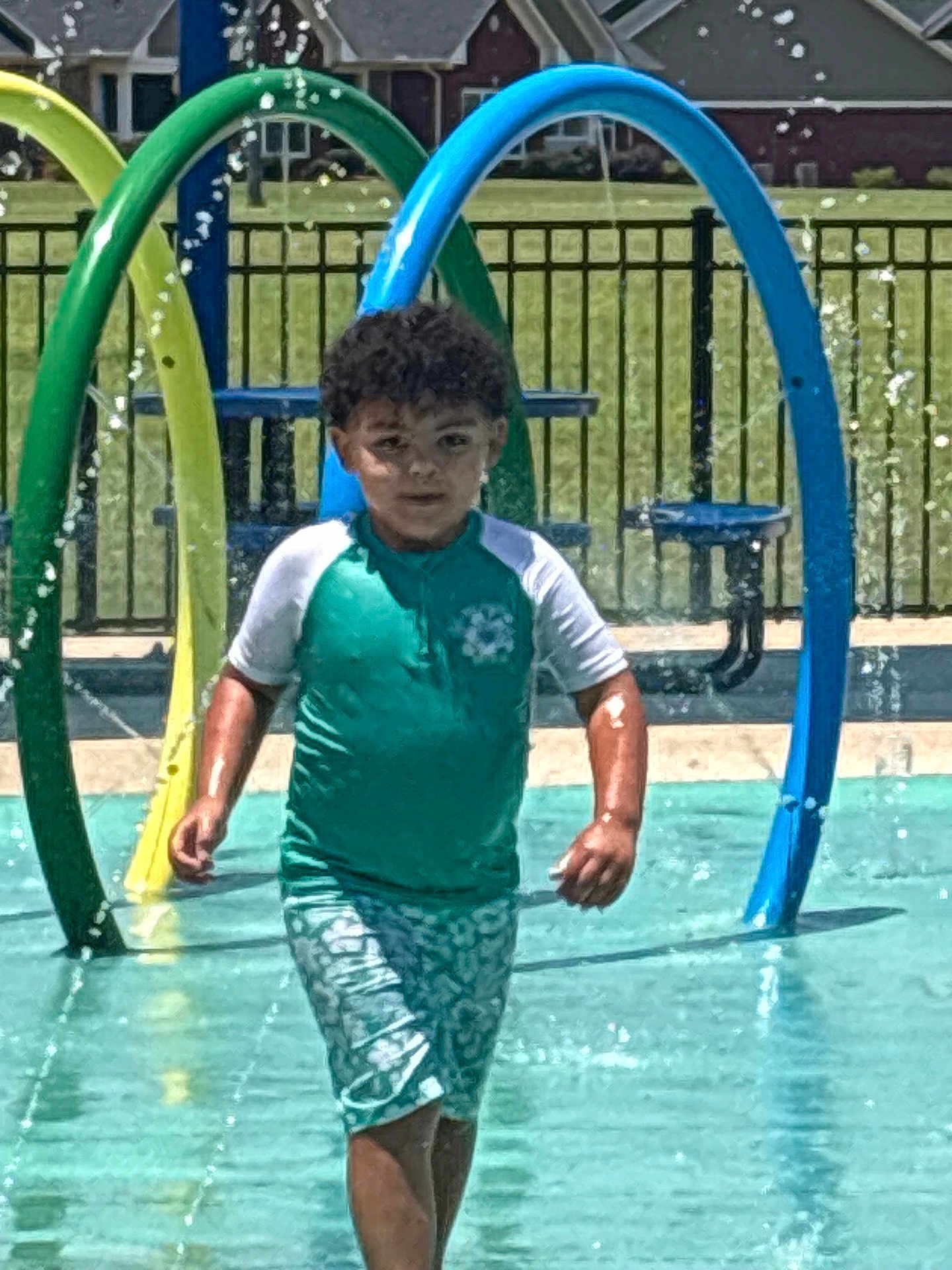 Joseph is registered to the contest to win money with this photo: child, water, splash_pad, water_spray, playground, summer, outdoor, water_park, boy, shorts, tshirt, curly_hair, fence, sunny, recreation, water_feature, activity, fun, daytime, park