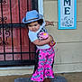 Bailey is registered to the contest to win money with this photo: child, toddler, cowboy_hat, pink_pants, sippy_cup, door, doormat, playful_pose, curly_hair, shirt, footwear, outdoor, person, smiling, curious, home_decor, wall, window, iron_grill, floor
