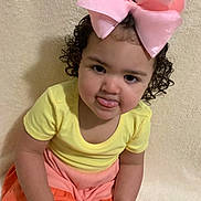 Bailey is registered to the contest to win money with this photo: toddler, child, curly_hair, pink_bow, yellow_top, orange_pants, playful, tongue_out, sitting, indoor, portrait, cute, happy, baby, person, casual_clothing, smiling, young_child, face, background