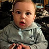 baby, child, indoor, hoodie, curious, wide_eyes, person, holding, small_object, casual_clothing, cute, portrait, seated, infant, expression, young_child, home, cozy, light, face