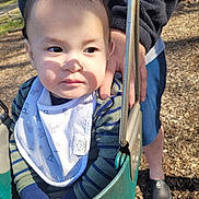 Zachary is registered to the contest to win money with this photo: adult_hand, bib, casual_clothing, child, curious, daylight, face, footwear, green_swing, hand, outdoor, person, playground, shadow, striped_clothing, sunlight, support, swing, toddler, wood_chips