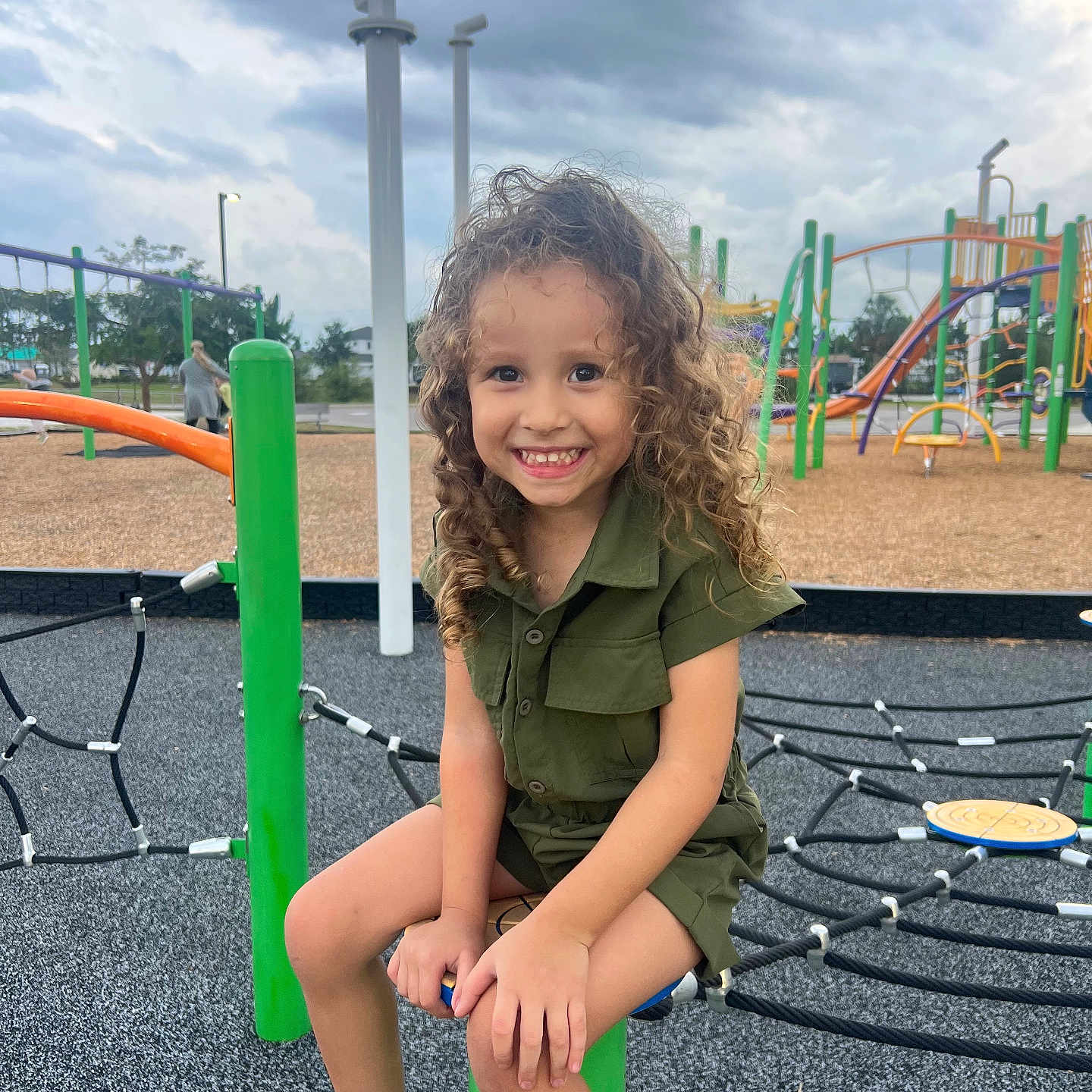 Zyli is registered to the contest to win money with this photo: active, casual_clothing, child, cloudy_sky, curly_hair, daylight, fun, green_outfit, green_post, happy, outdoor, park, person, play_equipment, playground, rope_climbing_structure, shoes, sitting, smiling, young