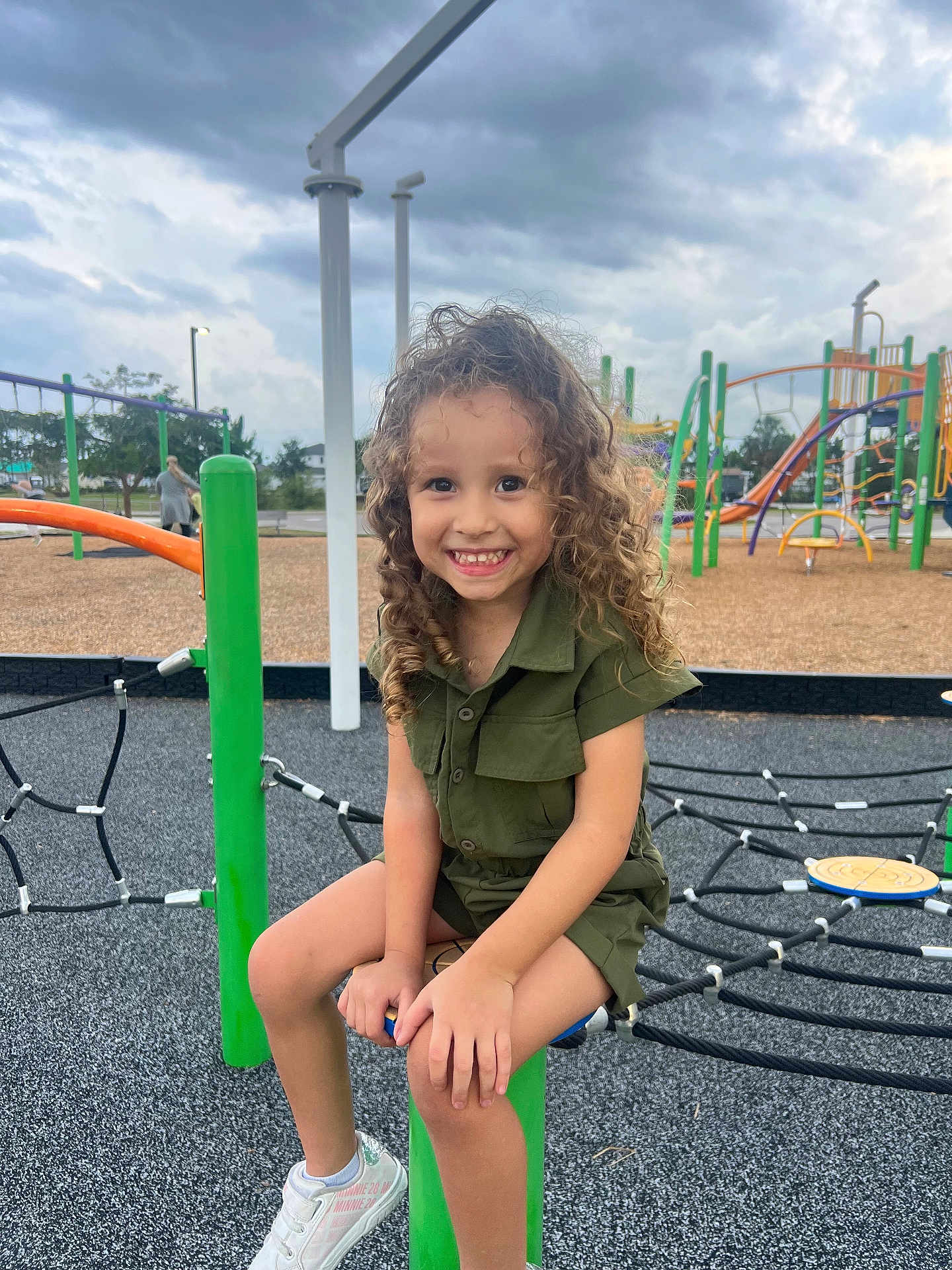 Zyli is registered to the contest to win money with this photo: child, curly_hair, green_outfit, playground, smiling, sitting, green_post, rope_climbing_structure, shoes, outdoor, cloudy_sky, happy, park, play_equipment, person, daylight, fun, casual_clothing, young, active