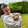 affection, animal, calm, casual_clothing, cute, daylight, dog, grass, greenery, hedge, holding, hoodie, nature, outdoor, pet, portrait, puppy, smile, woman, young_adult