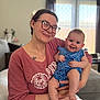 woman, baby, glasses, indoor, tattoo, smile, holding, sweatshirt, blue_dress, curtains, couch, natural_light, happy, person, portrait, child, home, casual_clothing, earrings, facial_expression