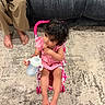 toddler, child, baby_bottle, pink_stroller, carpet, sofa, bare_feet, indoor, curly_hair, pink_dress, toy, sitting, person, floor, furniture, relaxed, home, young_child, casual, cute