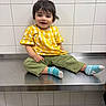 child, toddler, smiling, yellow_shirt, green_pants, blue_socks, sitting, metal_surface, white_tiles, indoor, cute, happy, portrait, clothing, person, young_child, casual, hair, face, feet