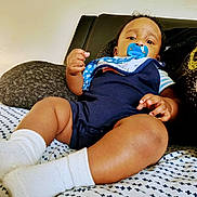 Mikko is registered to the contest to win money with this photo: baby, pacifier, onesie, white_socks, bedspread, pattern, infant, child, indoors, curly_hair, face, skin, hand, footwear, furniture, couch, comfort, resting, portrait, young_child