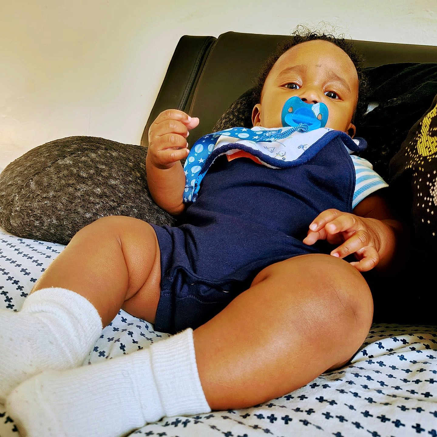 Mikko is registered to the contest to win money with this photo: baby, bedspread, child, comfort, couch, curly_hair, face, footwear, furniture, hand, indoors, infant, onesie, pacifier, pattern, portrait, resting, skin, white_socks, young_child