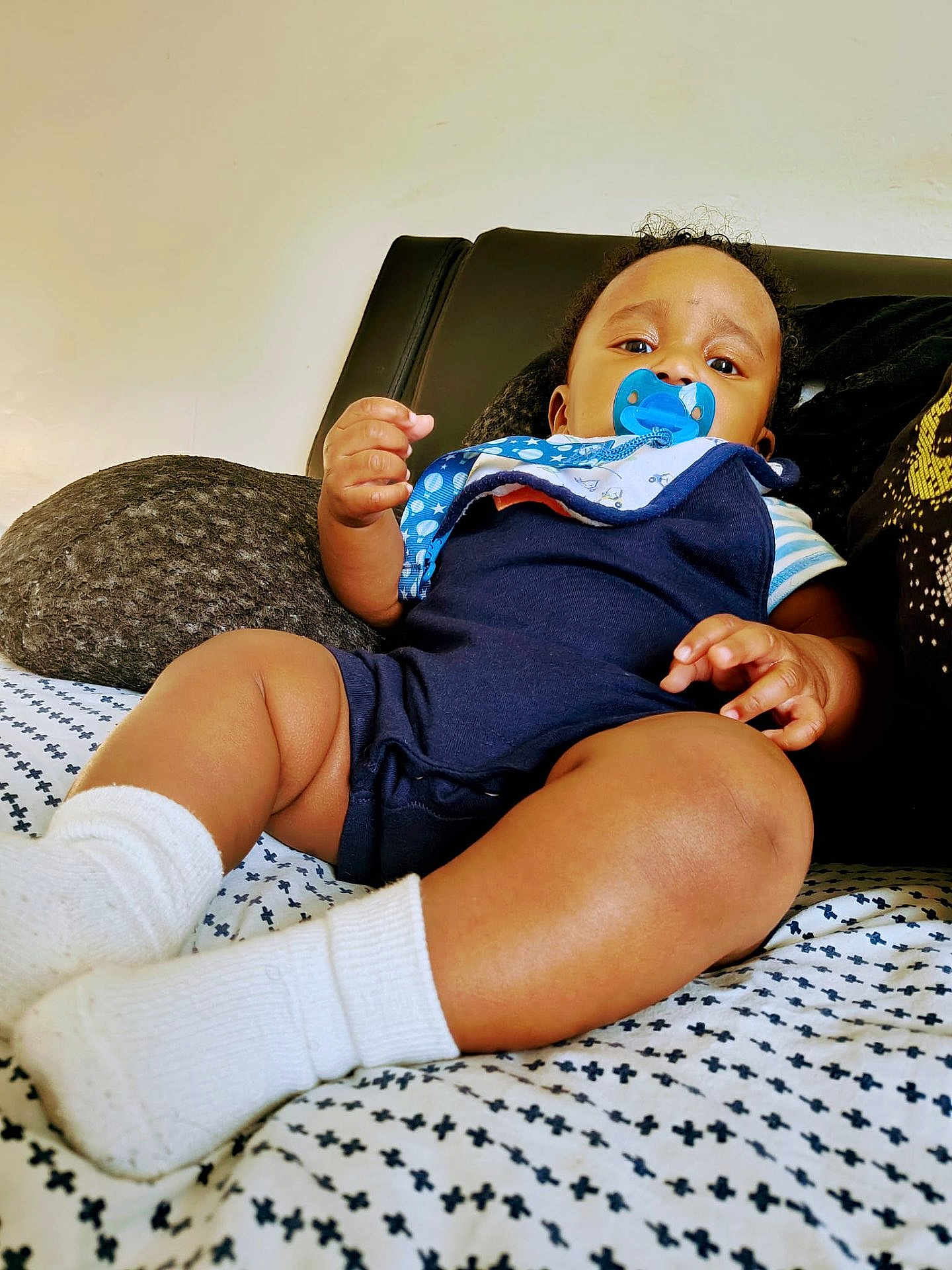 Mikko is registered to the contest to win money with this photo: baby, pacifier, onesie, white_socks, bedspread, pattern, infant, child, indoors, curly_hair, face, skin, hand, footwear, furniture, couch, comfort, resting, portrait, young_child