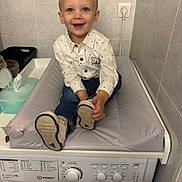 Tiago participe au concours pour gagner de l'argent avec cette photo : toddler, child, smiling, shirt, jeans, washing_machine, changing_pad, indoor, bathroom, tiles, footwear, happy, person, electric_outlet, plastic_container, laundry, cleaning, appliance, young_child, home