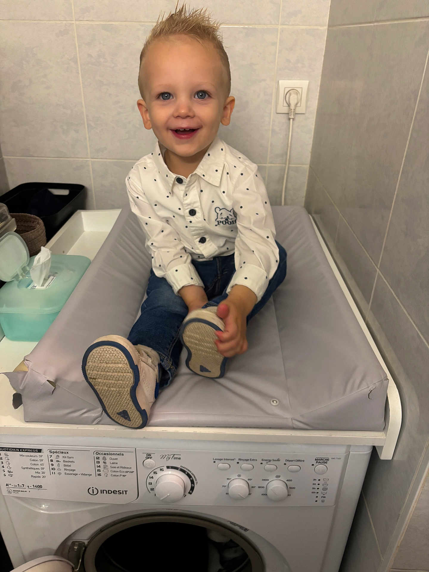Tiago participe au concours pour gagner de l'argent avec cette photo : toddler, child, smiling, shirt, jeans, washing_machine, changing_pad, indoor, bathroom, tiles, footwear, happy, person, electric_outlet, plastic_container, laundry, cleaning, appliance, young_child, home