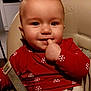 baby, child, high_chair, red_shirt, snowflake_pattern, smiling, finger_in_mouth, indoor, seatbelt, cute, face, human, young_child, holiday_theme, playful, clothing, person, portrait, cheerful, infant