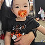 Athena is registered to the contest to win money with this photo: toddler, child, pacifier, headband, bow, halloween_shirt, adult_hand, smartwatch, nail_polish, living_room, carpet, toy_unicorn, toy, person, black_clothing, orange, purple, cute, indoor, playtime