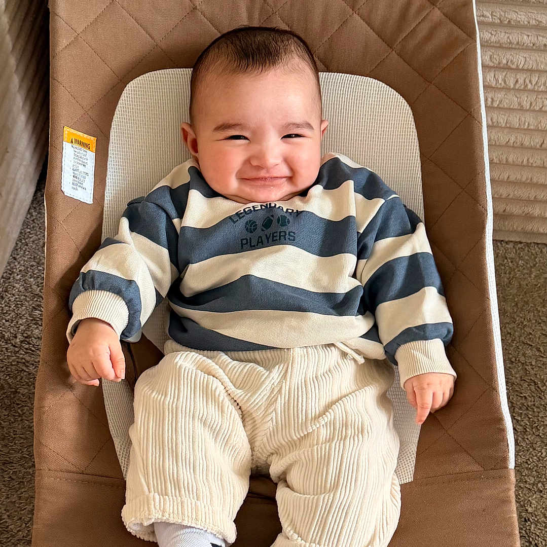 Ali joined the competition — help win amazing prizes! baby, smiling, striped_sweater, pants, socks, baby_rocker, indoor, carpet, face, person, child, happy, cute, infant, clothing, furniture, cozy, portrait, seated, relaxed