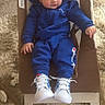 baby, child, blue_clothing, tracksuit, sneakers, baby_rocker, carpet, plush_toys, indoor, person, footwear, casual_wear, infant, cute, relaxed, soft_texture, floor, seated, young_child, cozy