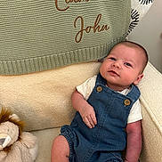 Liam John joined the competition — help win amazing prizes! baby, infant, denim_overalls, white_shirt, plush_toy, lion_plush, knitted_blanket, embroidered_text, cozy_chair, beige_fabric, smiling, cute, child, portrait, indoors, soft_lighting, relaxed, comfortable, young_child, person