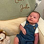 baby, infant, denim_overalls, white_shirt, plush_toy, lion_plush, knitted_blanket, embroidered_text, cozy_chair, beige_fabric, smiling, cute, child, portrait, indoors, soft_lighting, relaxed, comfortable, young_child, person