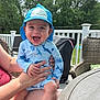 Rhys is registered to the contest to win money with this photo: baby, child, smiling, hat, blue_clothing, outdoor, sunlight, person, hand, lap, wicker_chair, table, greenery, trees, deck, happy, cute, infant, summer, shark_pattern