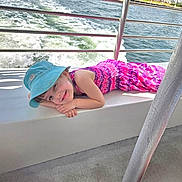 Myra is registered to the contest to win money with this photo: bench, blue_hat, boat, casual, child, concrete_floor, hand, happy, ocean, outdoor, palm_trees, pink_dress, portrait, rail, relaxing, smiling, summer, sunny, vacation, water