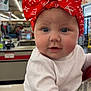 Kimberly is registered to the contest to win money with this photo: baby, child, headband, red_bow, white_clothing, indoor, store, fluorescent_lighting, face, person, cute, chubby_cheeks, holding, infant, portrait, shopping, background_people, expression, closeup, curious