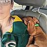 Teddy James is registered to the contest to win money with this photo: puppy, dog, brown_dog, sleeping, cuddling, lap, person, face, glasses, car, car_interior, seat, headrest, collar, green_collar, road_trip, pet, cute, portrait, jacket