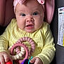 baby, infant, headband, pink_bow, yellow_sweater, car_seat, teething_toys, colorful, hands, cute, child, face, portrait, indoor, expression, holding, playful, soft_lighting, close_up, sitting