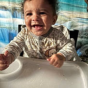 Kaysyn joined the competition — help win amazing prizes! baby, child, high_chair, smiling, curly_hair, messy_face, onesie, indoor, feeding_time, happy, face, hands, table, food_crumbs, background, colorful, person, portrait, cute, infant