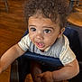 toddler, child, curly_hair, high_chair, indoor, wooden_floor, white_shoes, short_sleeve_shirt, cute, expression, person, furniture, table, cute_face, looking_up, casual_clothing, young_child, seat, curious, small_shoes