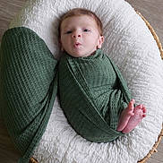 Owen joined the competition — help win amazing prizes! baby, infant, swaddled, green_wrap, basket, white_cushion, feet, face, wooden_floor, newborn, resting, curious_expression, cozy, textured_fabric, indoors, portrait, child, small_feet, wrapped, cute
