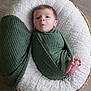 baby, infant, swaddled, green_wrap, basket, white_cushion, feet, face, wooden_floor, newborn, resting, curious_expression, cozy, textured_fabric, indoors, portrait, child, small_feet, wrapped, cute