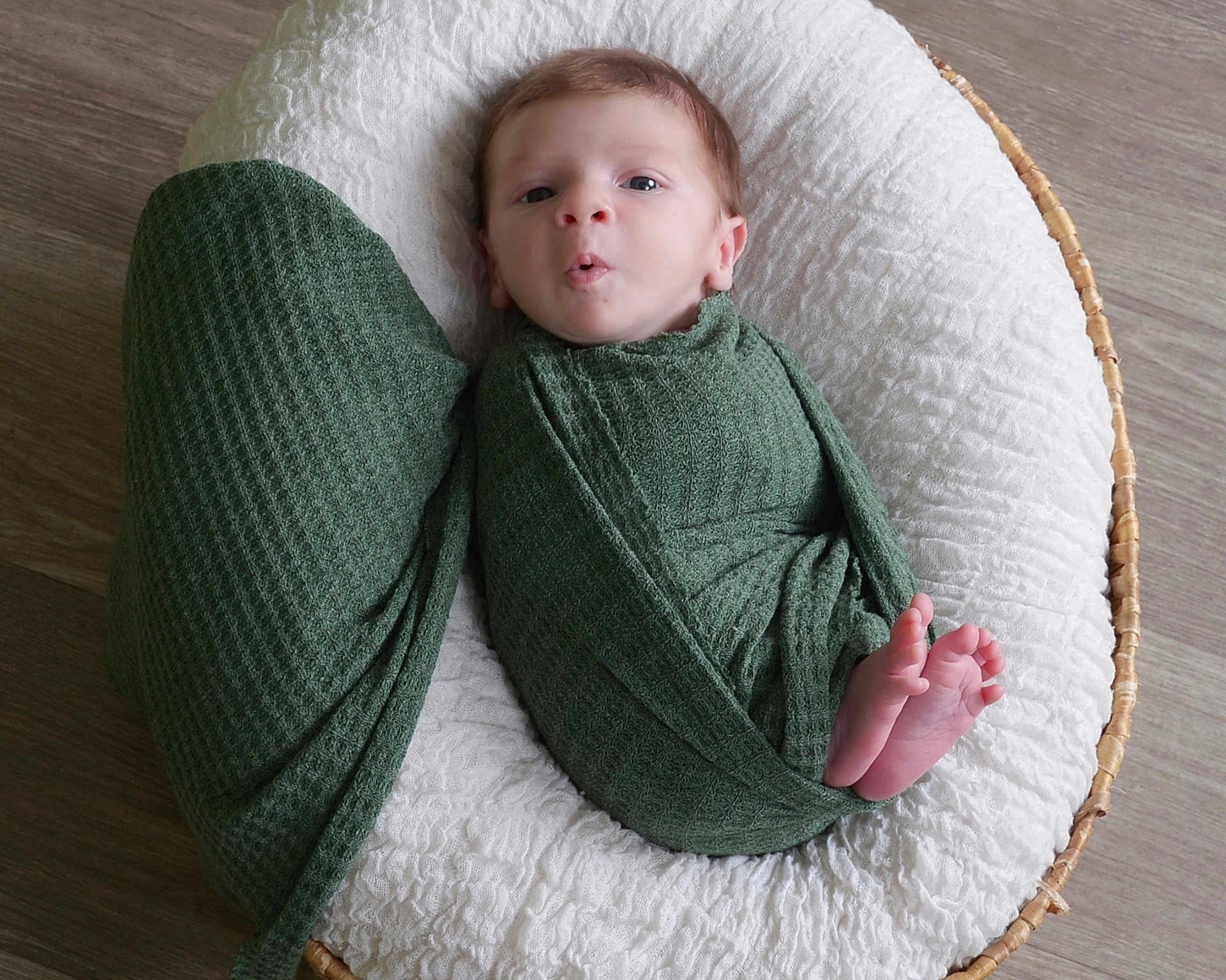 Owen joined the competition — help win amazing prizes! baby, infant, swaddled, green_wrap, basket, white_cushion, feet, face, wooden_floor, newborn, resting, curious_expression, cozy, textured_fabric, indoors, portrait, child, small_feet, wrapped, cute