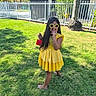 child, girl, yellow_dress, sunglasses, flower_sunglasses, red_purse, grass, lawn, tree, fence, backyard, sunlight, shadow, smiling, happy, outdoor, person, footwear, playful, summer