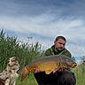 man, dog, fish, grass, reeds, sky, outdoor, nature, fishing, animal, person, water, catch, pet, greenery, clouds, wildlife, summer, hobby, leisure