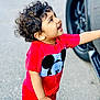 child, toddler, curly_hair, red_clothing, mickey_mouse, shirt, pants, smiling, happy, standing, outdoor, vehicle, tire, pavement, person, cute, expression, playful, casual, daylight