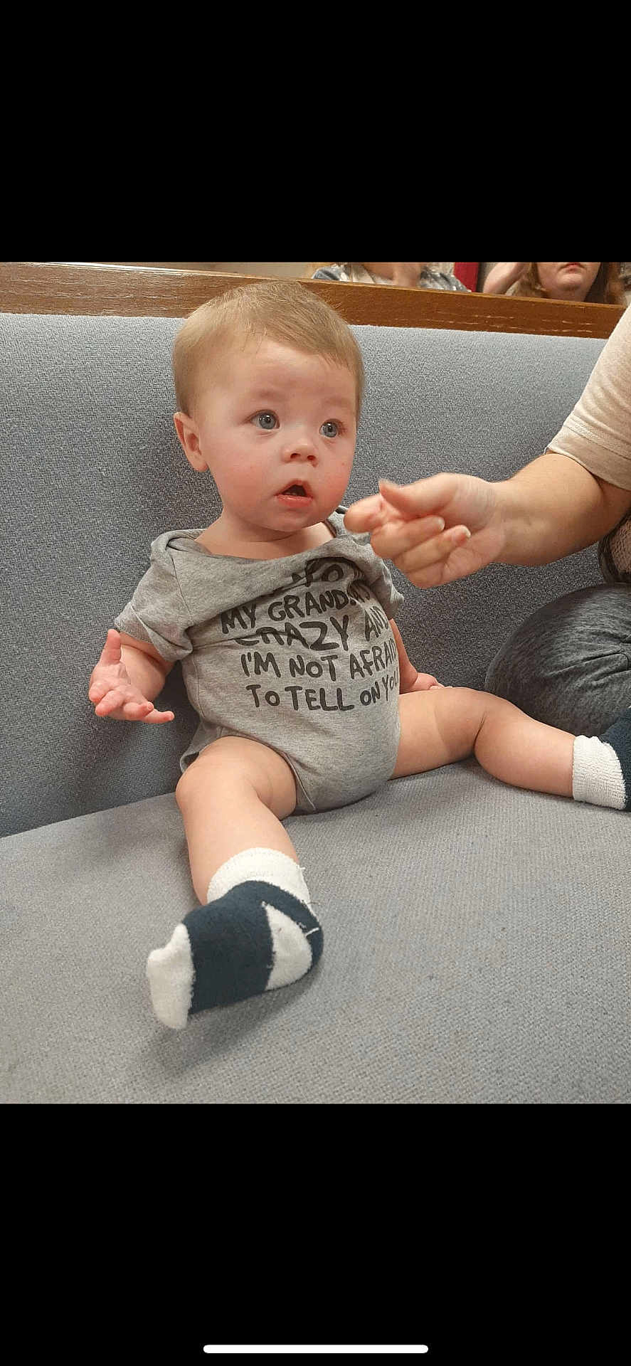 Levi joined the competition — help win amazing prizes! baby, child, infant, sitting, couch, gray_couch, onesie, clothing, sock, hand, person, indoor, expression, cute, curious, funny_text, casual, young, portrait, face